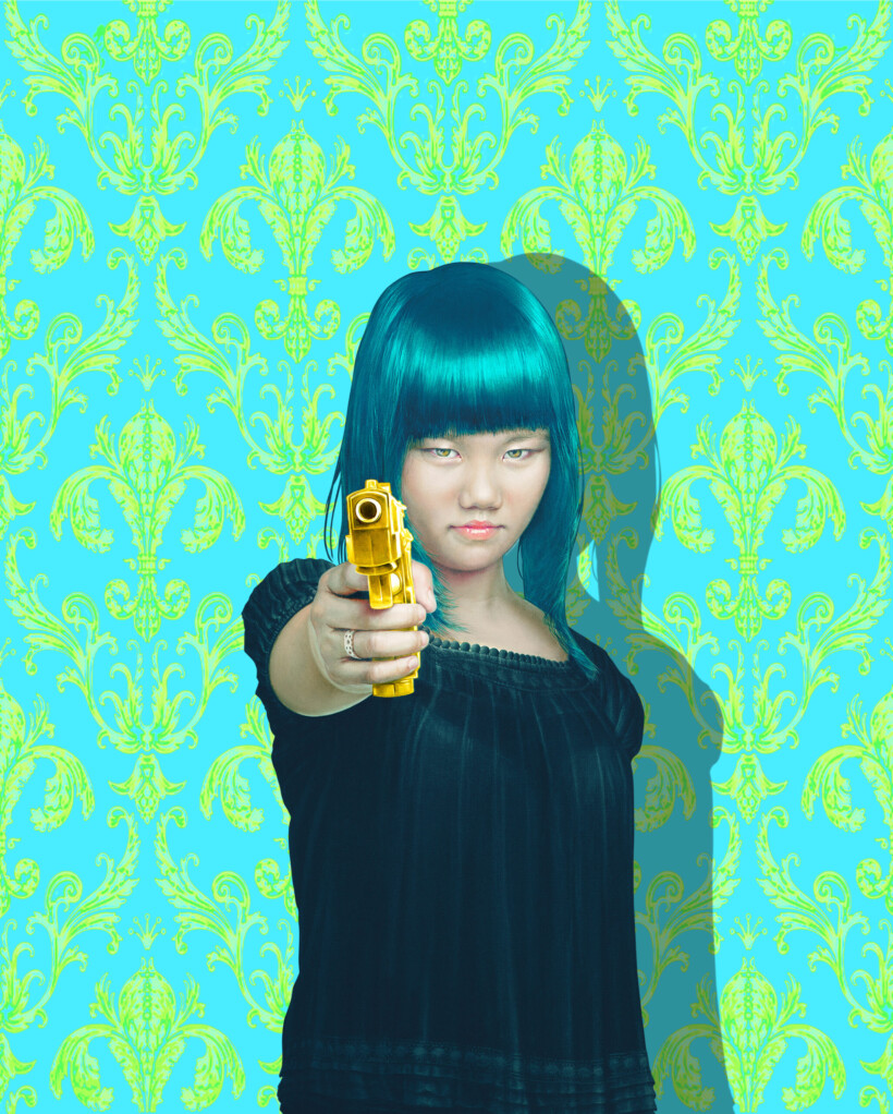 Una mujer con el pelo azul verdoso apuntando una pistola dorada hacia la cámara. Viste una blusa negra y el fondo es un papel tapiz azul turquesa con un patrón floral verde neón.